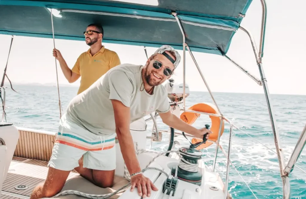 Zwei Männer genießen einen sonnigen Tag auf einer Segelyacht. Einer steuert das Ruder, der andere steht dahinter. Beide lächeln und tragen legere Sommerkleidung.