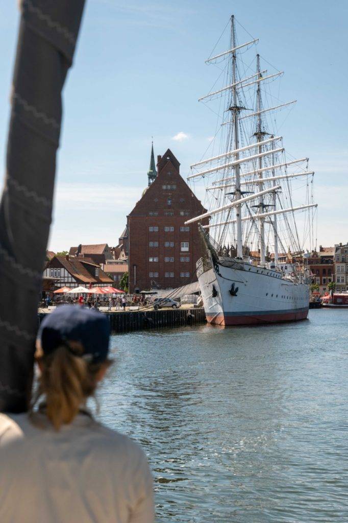 Eine Person betrachtet an einem sonnigen Tag an einer belebten Uferpromenade ein großes, vertäutes Segelboot, im Hintergrund sind historische Gebäude zu sehen, und denkt über einen Segelurlaub nach.