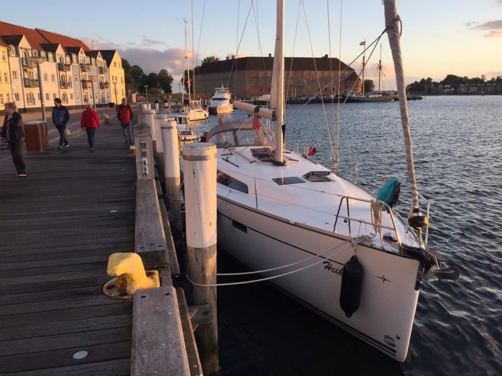 Ein Segelboot, das bei Sonnenuntergang an einem Holzsteg vor Anker liegt. Im Hintergrund sind flanierende Menschen und bunte Häuser zu sehen, die ihren Segelurlaub verbringen.