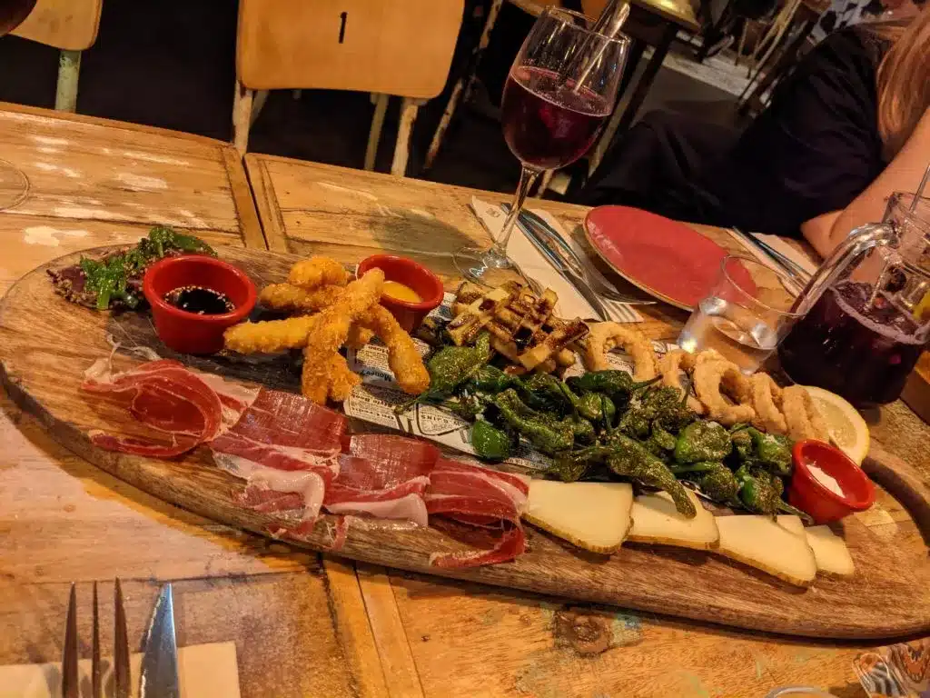 Auf einem Holztisch steht eine Wurstplatte mit verschiedenen Käsesorten, Wurstwaren, frittierten Calamari, gegrilltem Gemüse und Dip-Sauce, dazu gibt es während eines Segeltörns Gläser Rotwein.