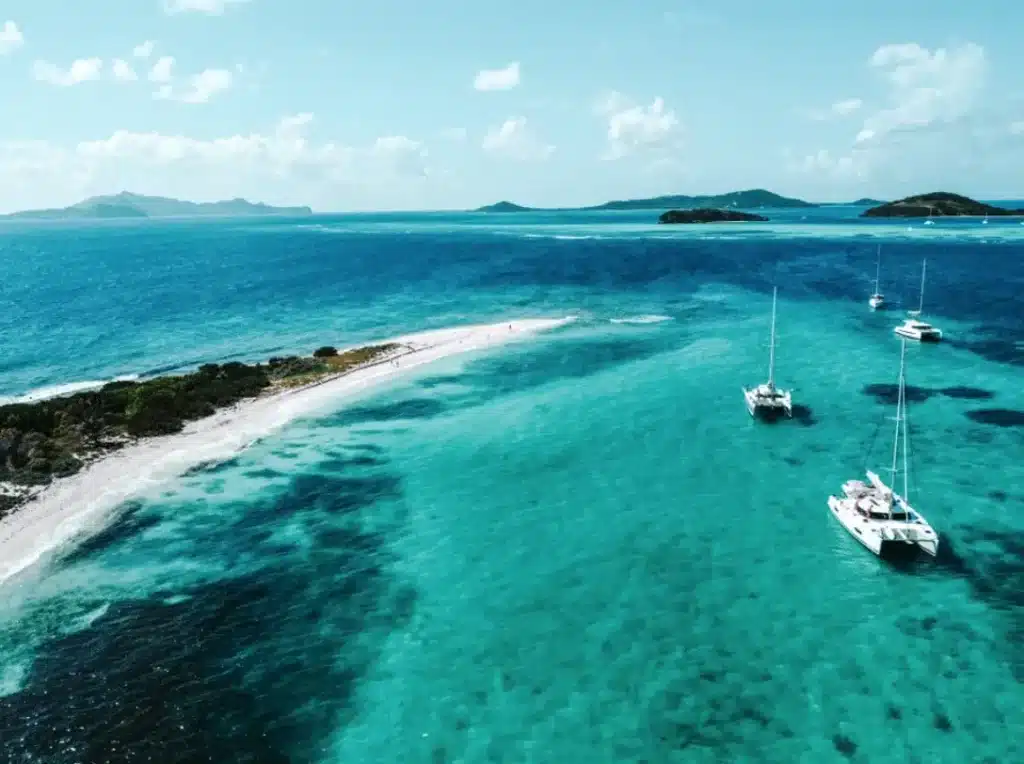 Luftaufnahme eines schmalen Sandstrands mit kristallklarem, türkisfarbenem Wasser, umgeben von mehreren Segelyachten vor einer üppig grünen Insel.