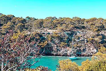 Ein malerischer Blick auf eine türkisfarbene Bucht, umgeben von üppigen grünen Bäumen und felsigen Klippen, mit einem kleinen Katamaran, der auf dem Wasser schwimmt. Darüber ein heller, klarer Himmel.