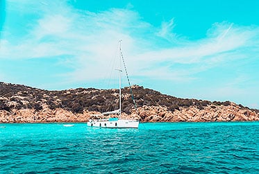 Ein weißer Katamaran schwimmt auf türkisfarbenem Wasser nahe einer felsigen Küste unter einem klaren blauen Himmel.