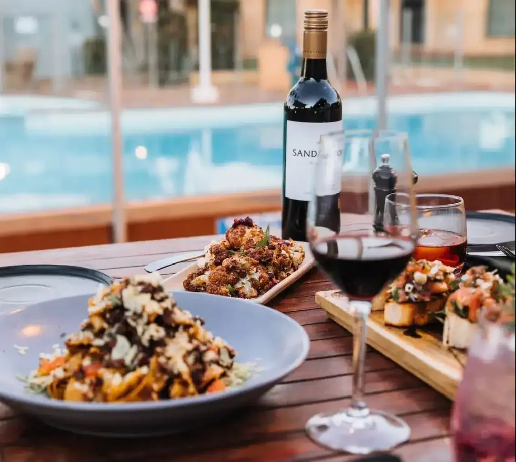 Ein gedeckter Tisch für ein Abendessen am Pool mit Gourmet-Gerichten, einer Flasche Rotwein und Weingläsern. Konzentrieren Sie sich auf die satten Farben und die Anordnung der Speisen, um eine einladende Atmosphäre zu schaffen, die an