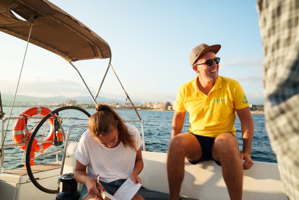 2 Personen auf einem Segelboot. Einer ist ein skipper im gelben Tshirt, das andere ist eine Frau, die segeln lernt. Der Skipper lacht und ist glücklich, sitzt auf der relig. Die Frau schaut runter und probiert was zu lesen. Die Sonne scheint und sie sind auf dem Meer auf dem Segelboot.