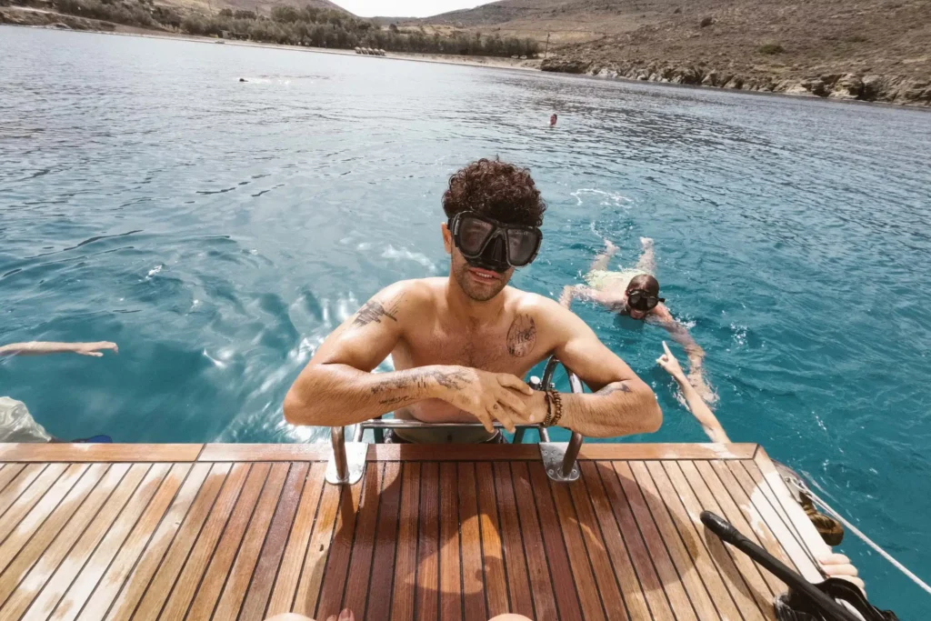 Eine Person mit Schwimmbrille steht auf einer Holzplattform am Rande des türkisfarbenen Wassers, im Hintergrund sind andere Schwimmer und vorbeigleitende Segelboote zu sehen. Die Plattform erstreckt sich bis ins Meer, umgeben von felsigen Hügeln unter einem teilweise bewölkten Himmel.