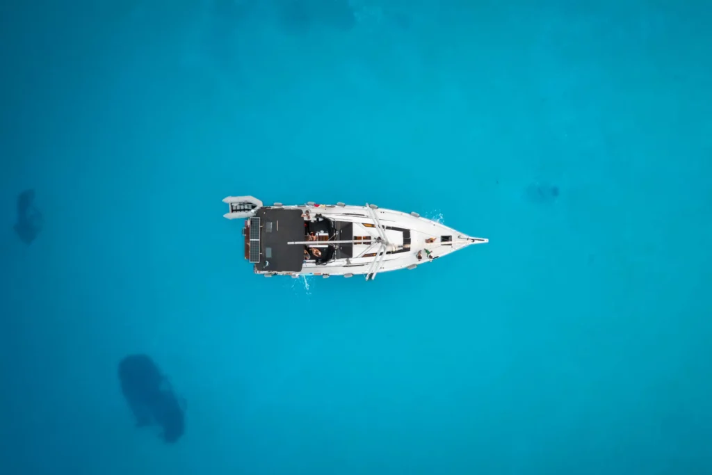 Luftaufnahme eines einzelnen weißen Segelboots, das in klarem, leuchtend blauem Wasser schwimmt. Das Boot befindet sich in der Bildmitte, umgeben von der unendlichen Weite des Meeres, und zeigt seine Ruhe und Abgeschiedenheit – eine heitere Segeltour auf ruhigem Wasser.