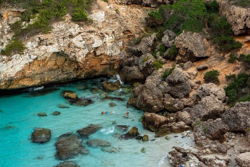 Eine versteckte Bucht mit türkisfarbenem Wasser, umgeben von schroffen Klippen und viel Grün, wo die Leute schwimmen und den abgeschiedenen Strandbereich von Segeltörn genießen.