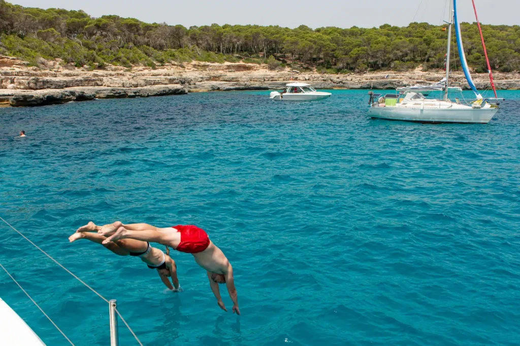 Ein Mann in roten Shorts springt von einer Segelyacht in das klare blaue Meer, mit anderen Booten in der Nähe und einer üppig grünen Küste im Hintergrund.