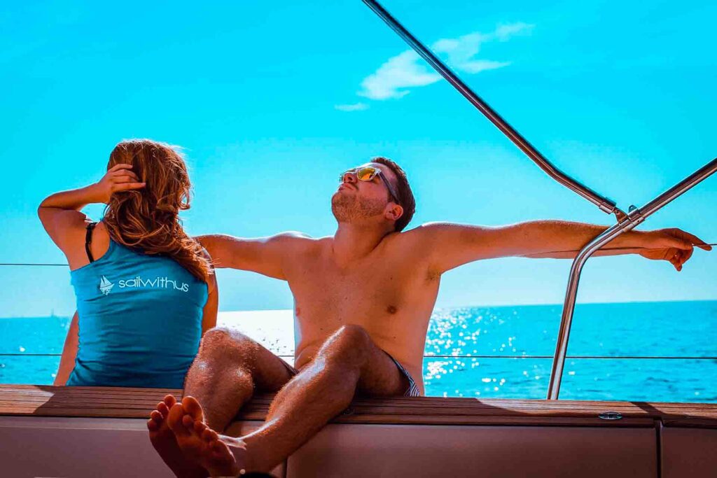 Ein Mann und eine Frau entspannen sich während ihres Segelurlaubs auf einer Yacht; der Mann streckt seine Arme auf der Reling aus, während er sich in der Sonne aalt, und die Frau sitzt neben ihm und bedeckt