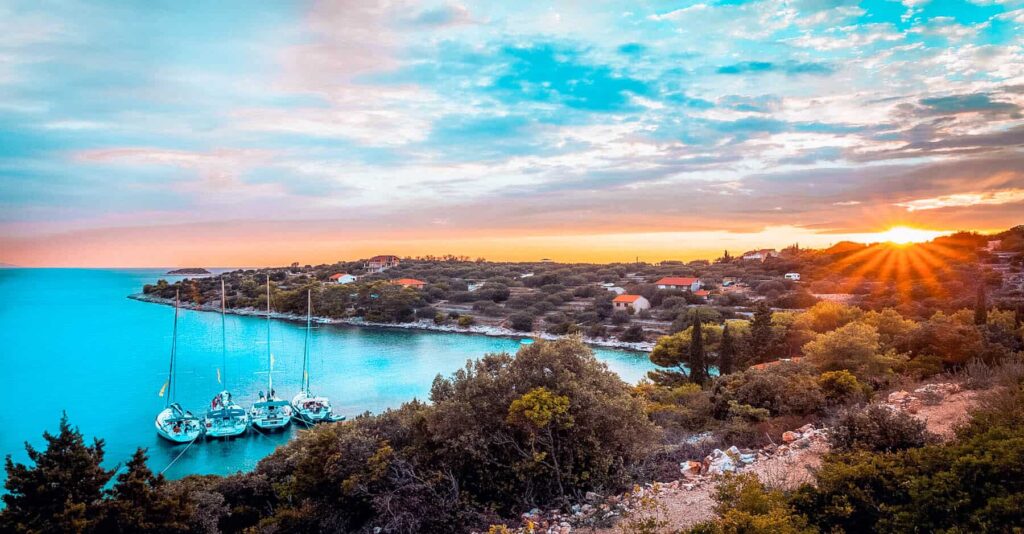 Sonnenuntergang über einer ruhigen Küstenlandschaft mit einem ruhigen blauen Meer mit vertäuten Segelyachten, umgeben von üppigem Grün und Häusern mit roten Dächern unter einem strahlenden Himmel.