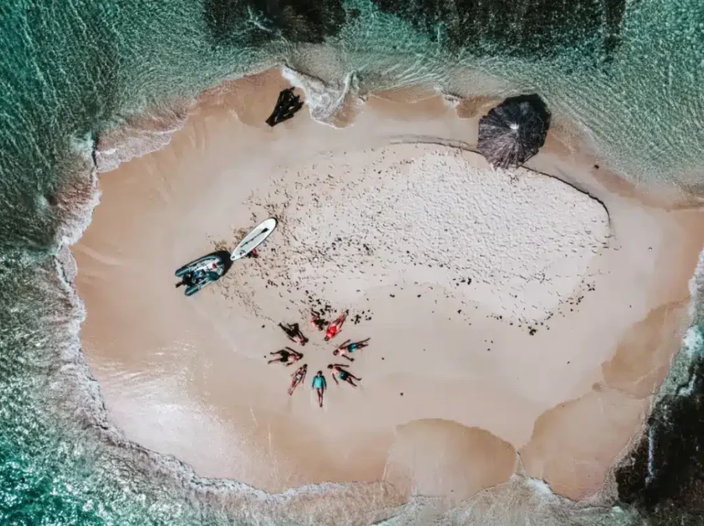 Luftaufnahme eines kleinen, abgelegenen Sandstrandes, umgeben von smaragdgrünem Wasser, mit mehreren Segelyachten in der Nähe und einer Gruppe von Menschen, die über den Sand verstreut sind.