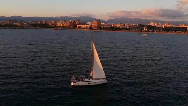 Ein Segelboot mit weißen Segeln gleitet bei Sonnenuntergang auf ruhigen Gewässern in der Nähe einer Küstenstadt. Die Skyline zeigt eine Mischung aus niedrigen und mittelhohen Gebäuden, mit Bergen im Hintergrund und einem teilweise bewölkten Himmel darüber.