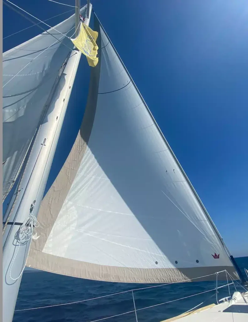 Die weißen Segel einer Segelyacht sind vollständig ausgefahren und sonnenbeschienen unter einem klaren, blauen Himmel. Die Perspektive ist von Bord des Schiffes, mit Blick nach oben zum Mast.