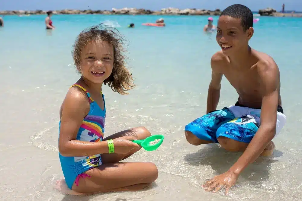 Ein junges Mädchen und ein Teenager lächeln und spielen im seichten Wasser des Strandes. Das Mädchen kniet mit einer grünen Sandschaufel, der Junge in blauen Badeshorts sieht ihr beim Segeltörn zu.