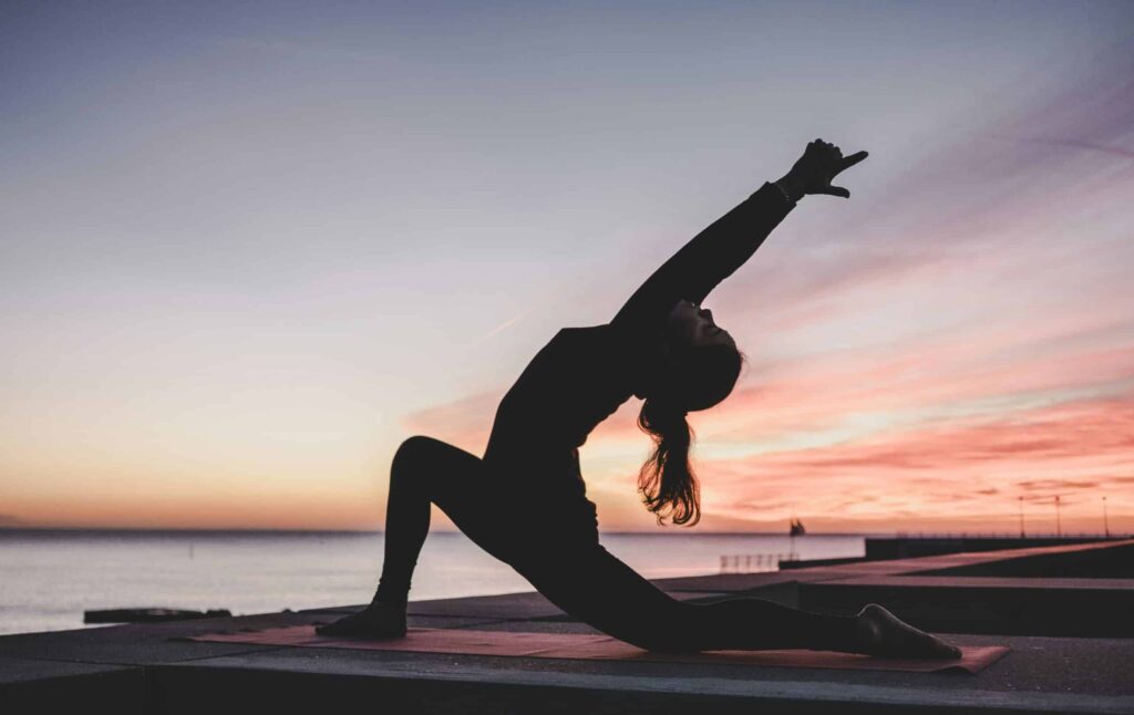 Eine Person als Silhouette macht in der Abenddämmerung eine Yoga-Pose auf einer Matte in der Nähe eines Gewässers. Im Hintergrund ist der Himmel in einem Farbverlauf von Orange nach Violett gehalten, der die untergehende Sonne anzeigt. In der Nähe schwimmt sanft eine majestätische Segelyacht und trägt zur ruhigen Atmosphäre dieses friedlichen Moments bei.