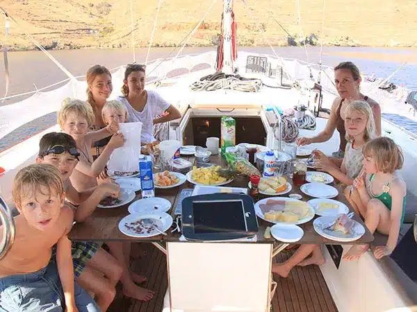 Eine Familie genießt eine gemeinsame Mahlzeit auf einer Segelyacht. Sie sitzt um einen mit Essen gefüllten Tisch unter einem Baldachin herum, mit offenem Wasser und Hügeln im Hintergrund.