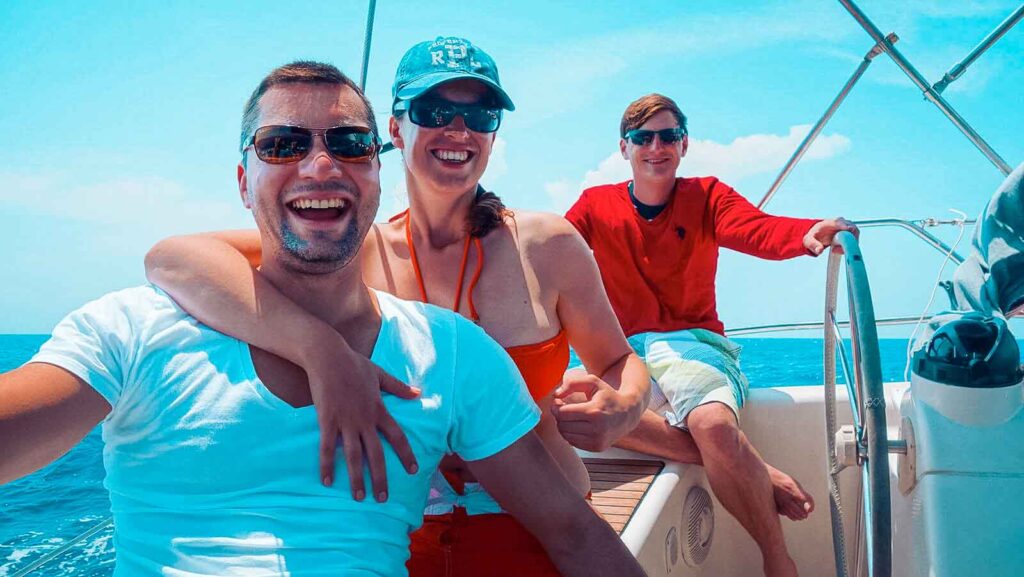 Drei Freunde genießen einen sonnigen Tag auf einer Segelyacht mit einem klaren blauen Himmel und dem Meer im Hintergrund. Zwei Männer und eine Frau lächeln und sitzen gemütlich da.